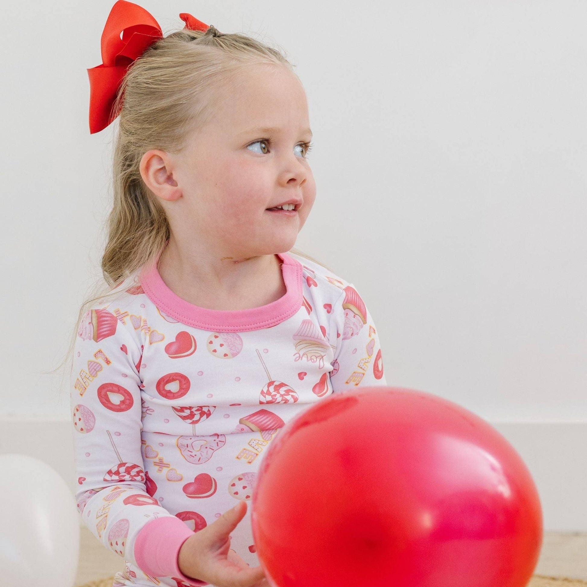 Child wearing pajamas with a red bow in hair, holding a red balloon against a white background