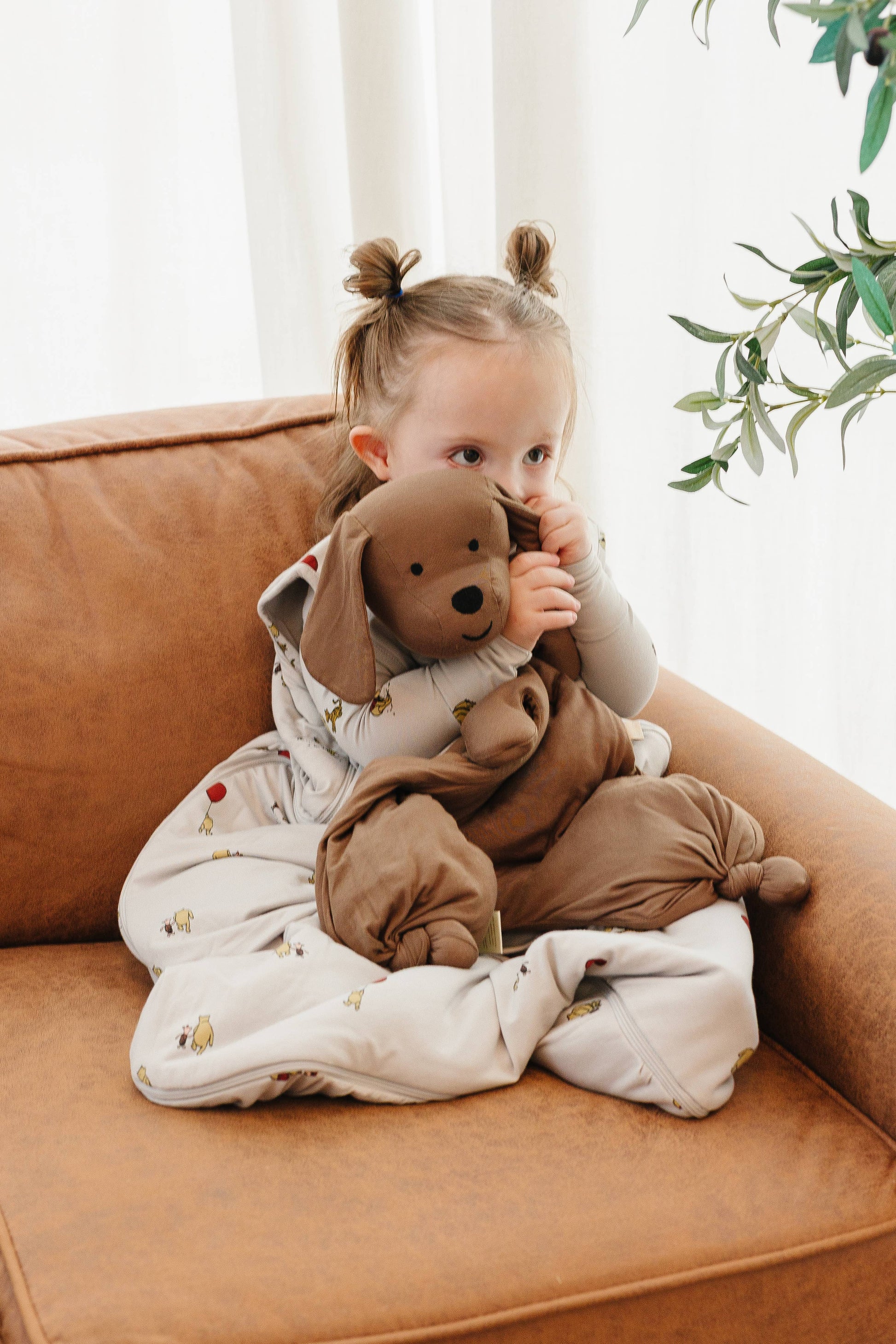 Child sitting on a couch holding a brown plush toy, wrapped in a white blanket with gold patterns.