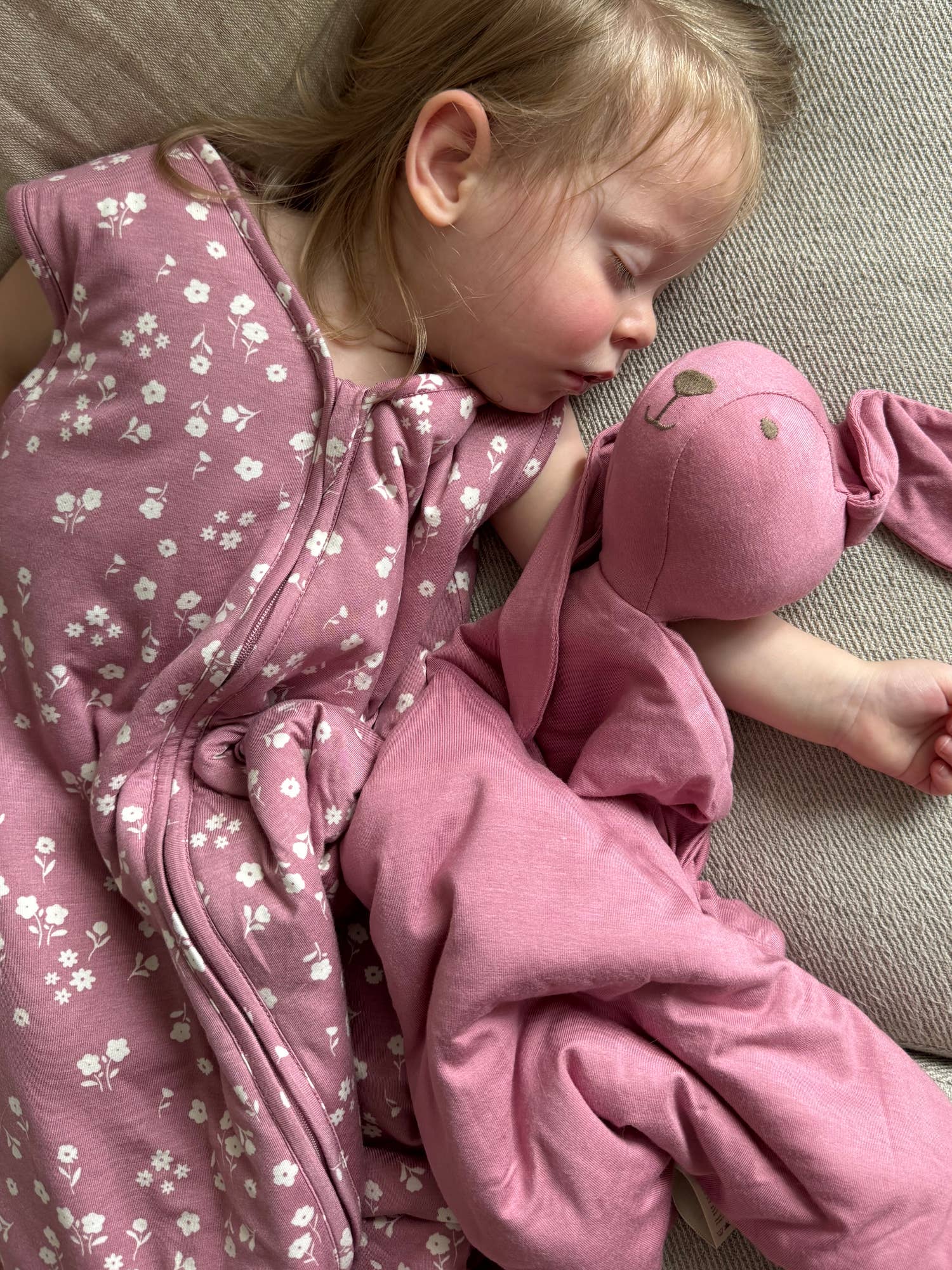 Child in a pink floral sleep sack lying on a couch with a pink plush lovey.
