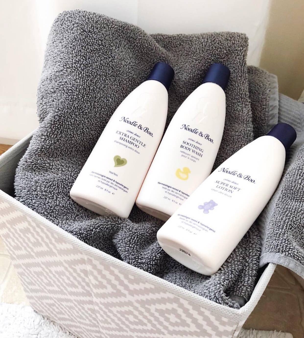 Three bottles of Noodle & Boo baby products on a gray towel inside a white basket.